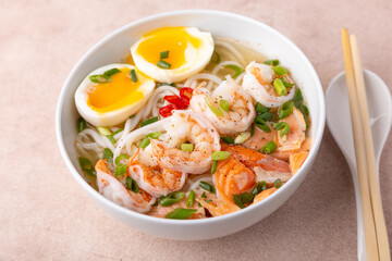 Asian dish with broth, noodles, tiger shrimp, fish, egg and green onions. Traditional Asian soup in a white deep plate. Selective focus, close-up.