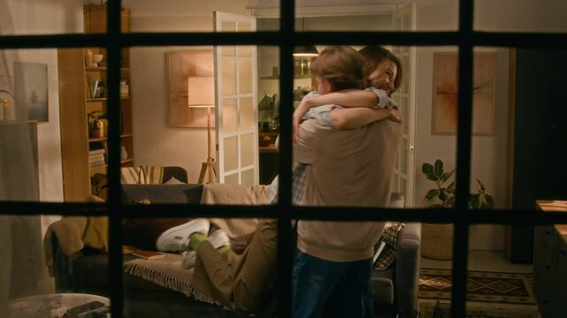 Wide shot, seen through window, of ecstatic young Caucasian man with laptop computer running into living room, sharing happy news with girlfriend of getting new job, both cheering and hugging