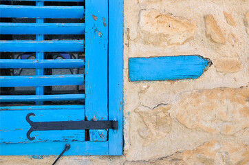 Rustic blue wooden window shutter on a textured stone wall, showcasing Mediterranean charm. The vibrant color contrasts with the rough stone, symbolizing tradition, architecture, and coastal vibes.
