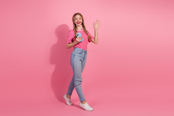 Young woman in casual summer attire holding smartphone against pink backdrop, exuding confidence and charm with a welcoming gesture.