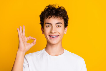 Smiling teenage boy with braces wearing a white t-shirt and making an OK gesture, standing against...