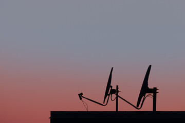 Two satellite dishes (or parabolic antenna), located on the roof of a residential building, used for television and telecommunication. Dusk sky on the background, with copy-space.