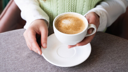 Close-up of woman's hands holding a white cup of hot coffee with creamy foam on a saucer in a cozy cafe. Coffee break, love for caffeine, pleasure of enjoying a warm beverage, cafeteria, coffee shop.