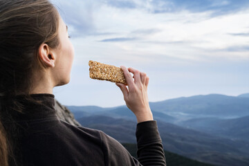 Back view of woman traveler eating a grain muesli cereal bar snack on the hike in the mountains....