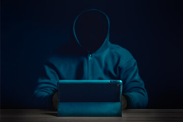 A mysterious figure in a dark hoodie uses a tablet in a dimly lit setting, representing cybercrime, hacking, and data security.