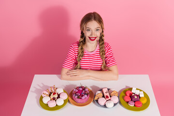 Cheerful young woman with braids and red lipstick sitting by desserts against pink background