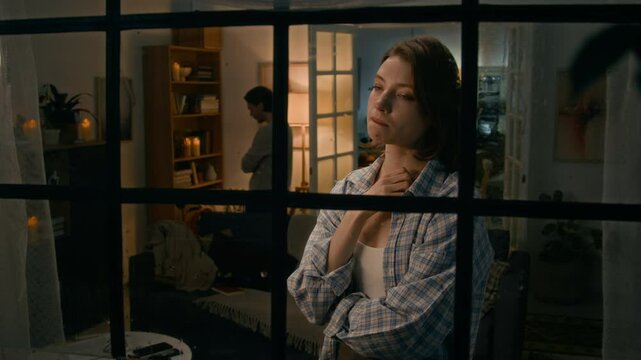 Medium shot of tearful young Caucasian girl standing by window during lighting storm and rain, crying after quarreling with boyfriend, man pacing around living room in background
