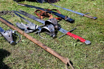 A close up on a set of medieval weapons and accessories, including swords, hammers, pikes, targes, furs, bucklers and drums laying on the grass seen on a sunny summer day during a historic fair