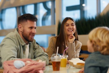 Adorable mother laughing out loud while her husband and son joking together during family lunch at food court in shopping mall, focus on parents
