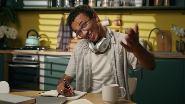 African American man ethnic guy high school student teen freelancer write homework task studying learning writing notebook at home kitchen looking at camera smiling male welcome hand invite gesture