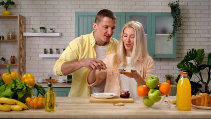 Couple of man and woman in the morning at home in the kitchen at table preparing breakfast, talking smiling, man helping woman make toasts with jam.