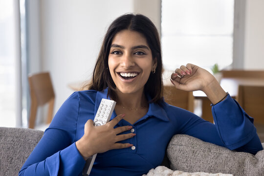 Young Indian female sitting comfortably on couch at home, holding remote control, smiling, feels excited, thrilled and pleasantly surprised while watching TV show or new series of favourite program