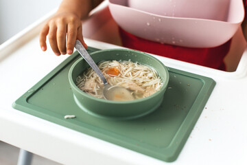 Close-up of a child's hand holding a spoon in a bowl of homemade chicken noodle soup. The child is...