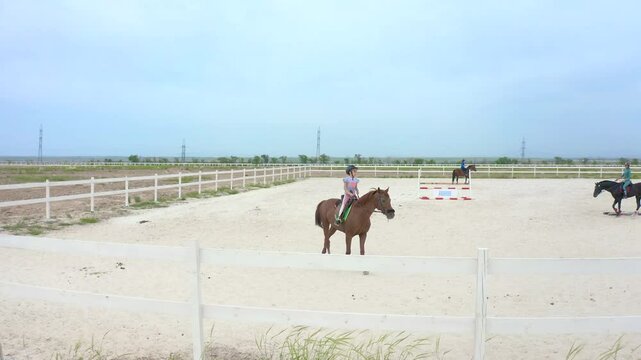 Horses and riders participate in equestrian sports on a sandy arena with jumps, surrounded by a white fence under a cloudy sky.