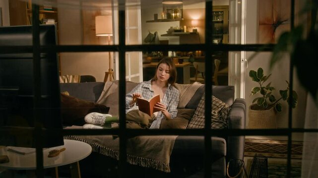 Full panning shot, observed by stranger or stalker through window, of young Caucasian girl relaxing alone in evening in cozy apartment, reading book on couch