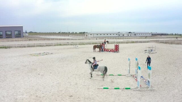 Aerial view of a horse riding arena, featuring equestrian practice with children and trainers engaging in jumping exercises in a sandy environment.