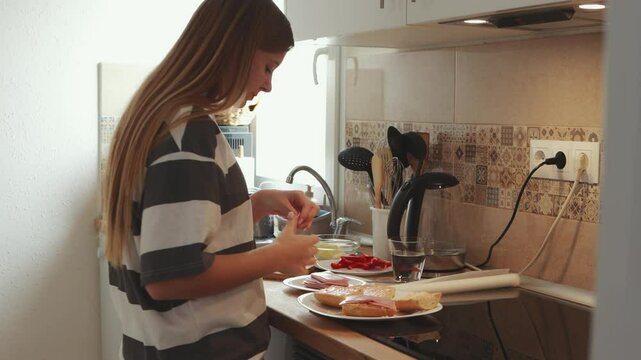 Young blonde teenager creating ham sandwich with red bell peppers on wooden cutting board, standing in bright contemporary white kitchen, wearing casual clothing