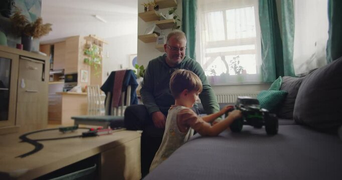Father smiling while his son plays with a green toy truck on a sofa, in a warm and cozy family living room with wooden furniture and a vibrant interior