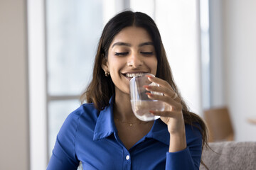 Attractive Indian woman holding glass of still or mineral water seated on sofa at home, making sip,...