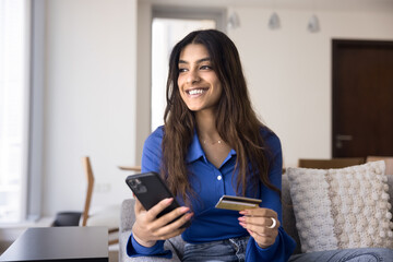 Fototapeta premium Young Indian woman holds card and phone buying via marketplace application, smile, sit on sofa at home, enjoy easy, instant money transfer through mobile app. E-shopping client purchasing on internet