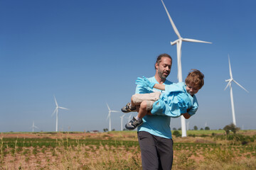A happy father carries his smiling son in the air with a windmill and a sky with clouds in the background.