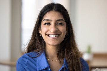 Head shot of beautiful young Indian woman sitting alone on sofa at home, posing for camera exude optimism, positive mood. Generation Z female, profile picture, portrait of virtual meeting participant