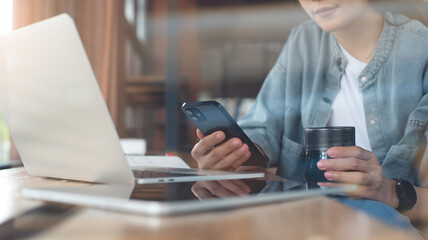 Fototapeta premium Asian business woman drinking coffee and using mobile phone during working on laptop computer at coffee shop. Freelancer online working, using smartphone surfing the internet at cafe