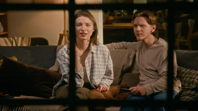 Medium full shot, seen from outside through window pane, of young Caucasian married couple and pet dog resting together on couch at night, watching horror movie, girl getting startled and screaming