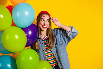Smiling young woman in casual denim outfit holding colorful balloons against vibrant yellow...