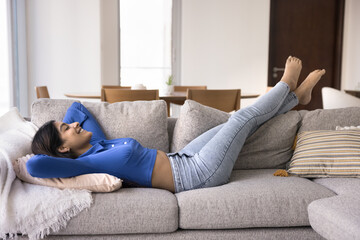 Side full-length view peaceful young Indian woman put hands behind head lying barefoot on sofa with...