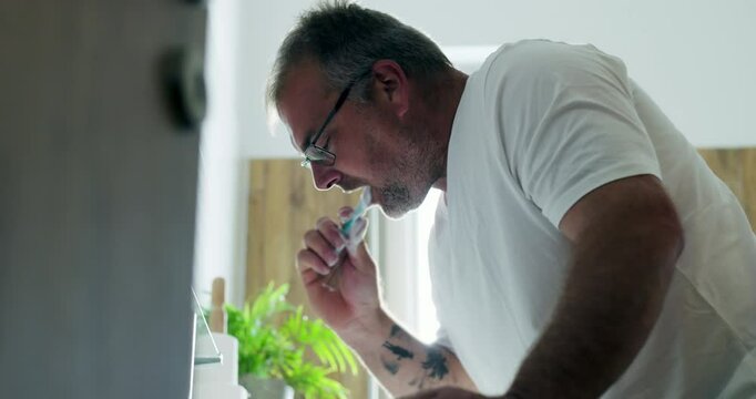 Man brushing teeth over sink, leaning slightly forward, focusing on self-care and oral hygiene as part of morning routine in clean and modern bathroom space
