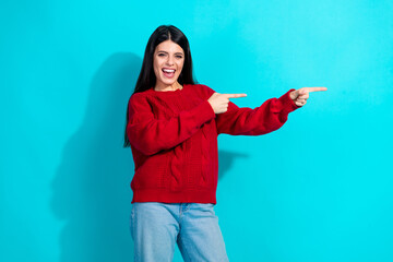 Cheerful young woman in a red sweater pointing to the side against a bright teal background,...