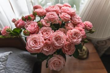 Obraz premium Top view of a large glass vase with a bouquet of pink roses on a bedside table by the window.
