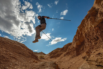 An extreme traveler jumps off the edge of a cliff with trekking poles against the backdrop of a...