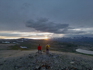 Two tourists in bright clothes stand during the golden hour with a view of the fiery sunset and snowy mountains. A photo from a quadcopter with a rear view.