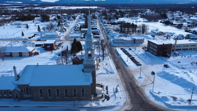 Winter drone approach toward the steeple of the Saint‑Nom‑de‑Marie Catholic church in the snow‑covered village of Sayabec, located in Quebec&rsquo;s Matap&eacute;dia Valley