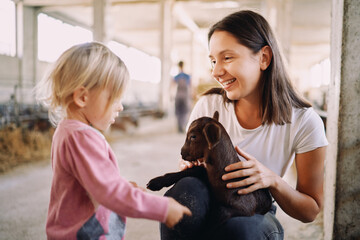 Little girl touches with her fingers a goatling in her mother arms on a farm