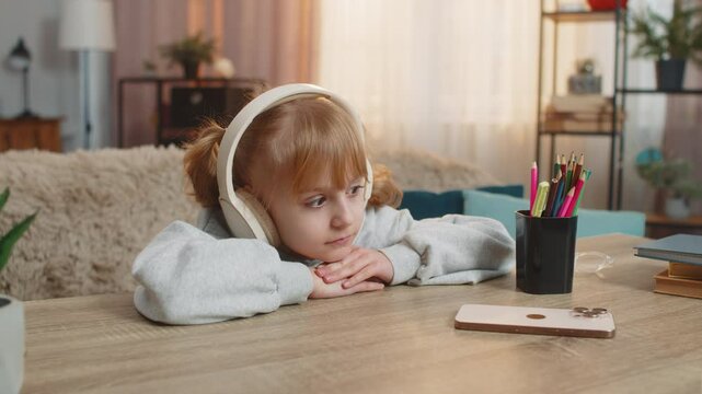 Young girl lying on table listening to an audiobook with headphones, reacting with excitement, surprise. Caucasian child smiles joyfully, wondering what happens next in story, enjoys adventure at home