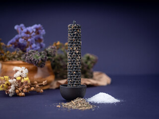 A magical black candle made with his own hands from wax wax with herbs lies on a purple background, dry herbs scattered. Magic to purify aura and energy