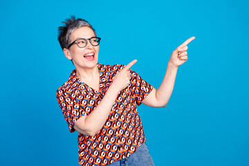 Cheerful mature woman with glasses pointing excitedly against a bright blue background in stylish...
