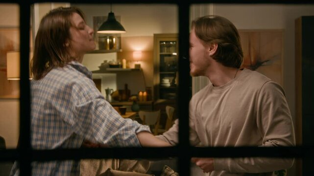 Medium shot of cheerful young Caucasian girl and guy enjoying dancing together to energetic music in living room, while having fun or celebrating happy event, observed through window pane