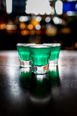 Three vibrant green shot glasses are arranged on a wooden table, softly illuminated against a blurred background of lights, creating a lively bar atmosphere.