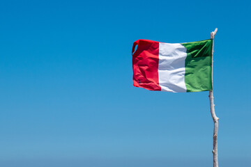 La bandiera italiana sventola al vento, attaccata ad un ramo bianco, sullo sfondo il cielo blu