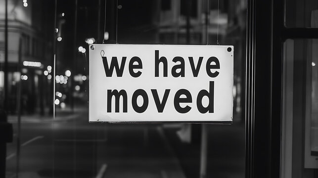 A monochrome image of a sign that reads "we have moved" hanging in a window, possibly indicating a business or residence has relocated to a new location.