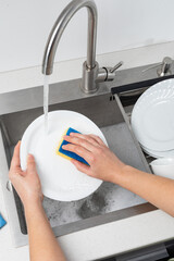 Housewife washing dishes in kitchen sink with sponge