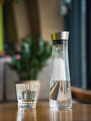 Stylish carafe filled with clear water stands next to short glass on polished wooden table. Background features greenery. Glass water pitcher and old fashioned rocks sit on wooden surface in cafe.