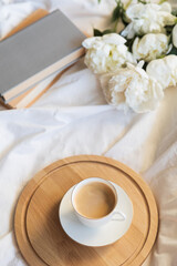 Coffee and peonies on a sunny cozy morning. Freshly brewed coffee cup rests on a wooden tray beside book and white peony flowers. A serene and cozy start to the day, soft natural light, calm ambiance