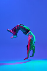 Dynamic image of flexible young guy in jeans and tank top dancing break dance against gradient blue purple background in neon light. Concept of dance, street style, creativity, youth culture, energy