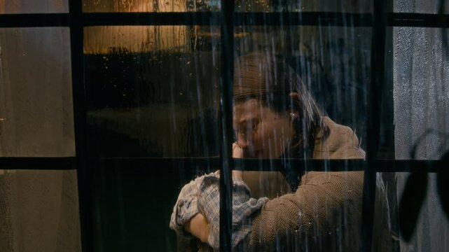 Medium closeup of young Caucasian woman with shawl around shoulders sitting at home on rainy night, crying in despair at relationship breakup while arguing on mobile phone while looking out of window