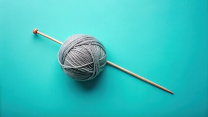 A ball of soft gray yarn rests beside a single knitting needle on a vibrant teal background, ready for a creative project